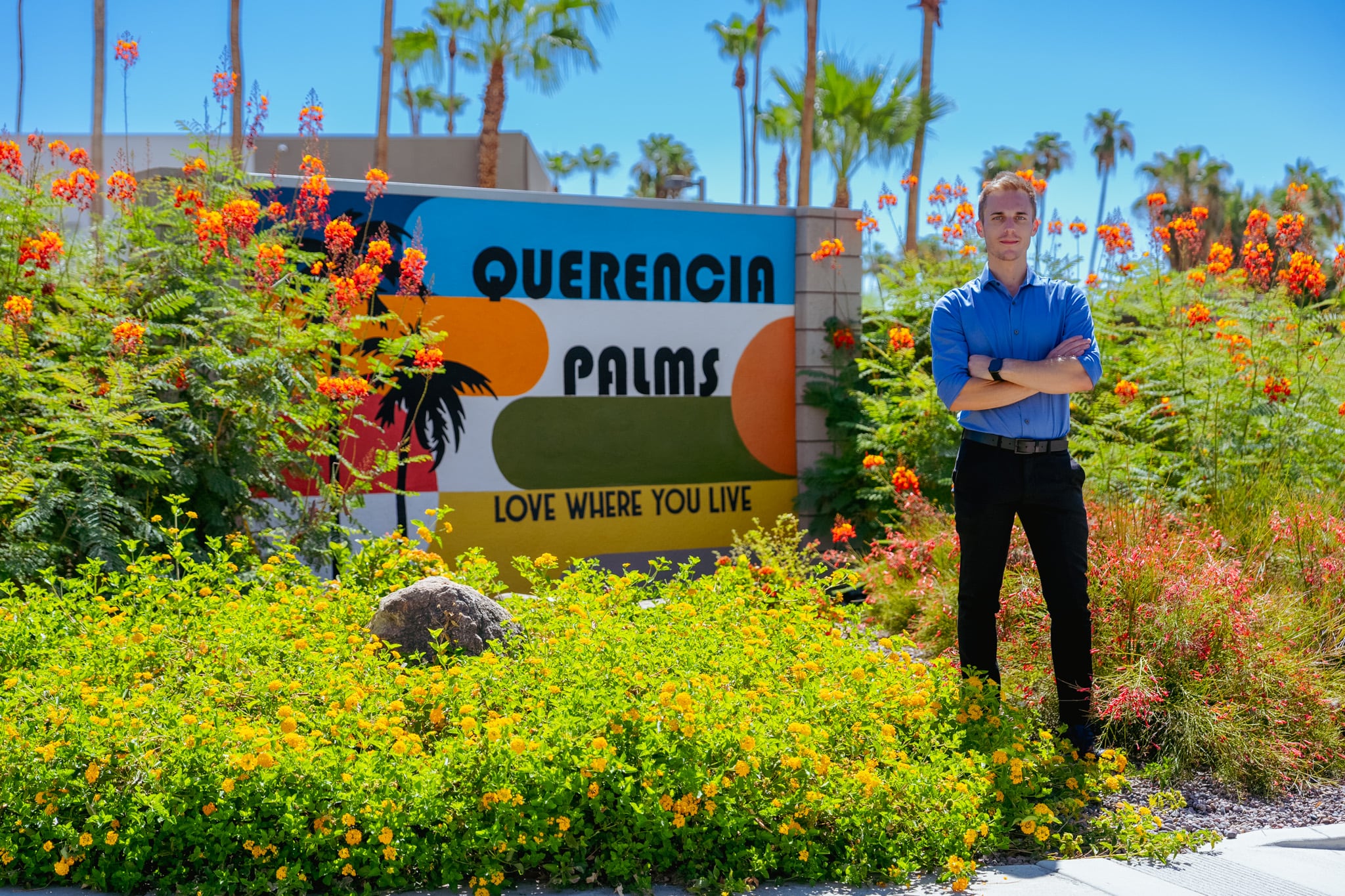 David Weis standing in front of the Querencia Palms mural in Palm Springs, surrounded by orange and yellow lantana in bloom