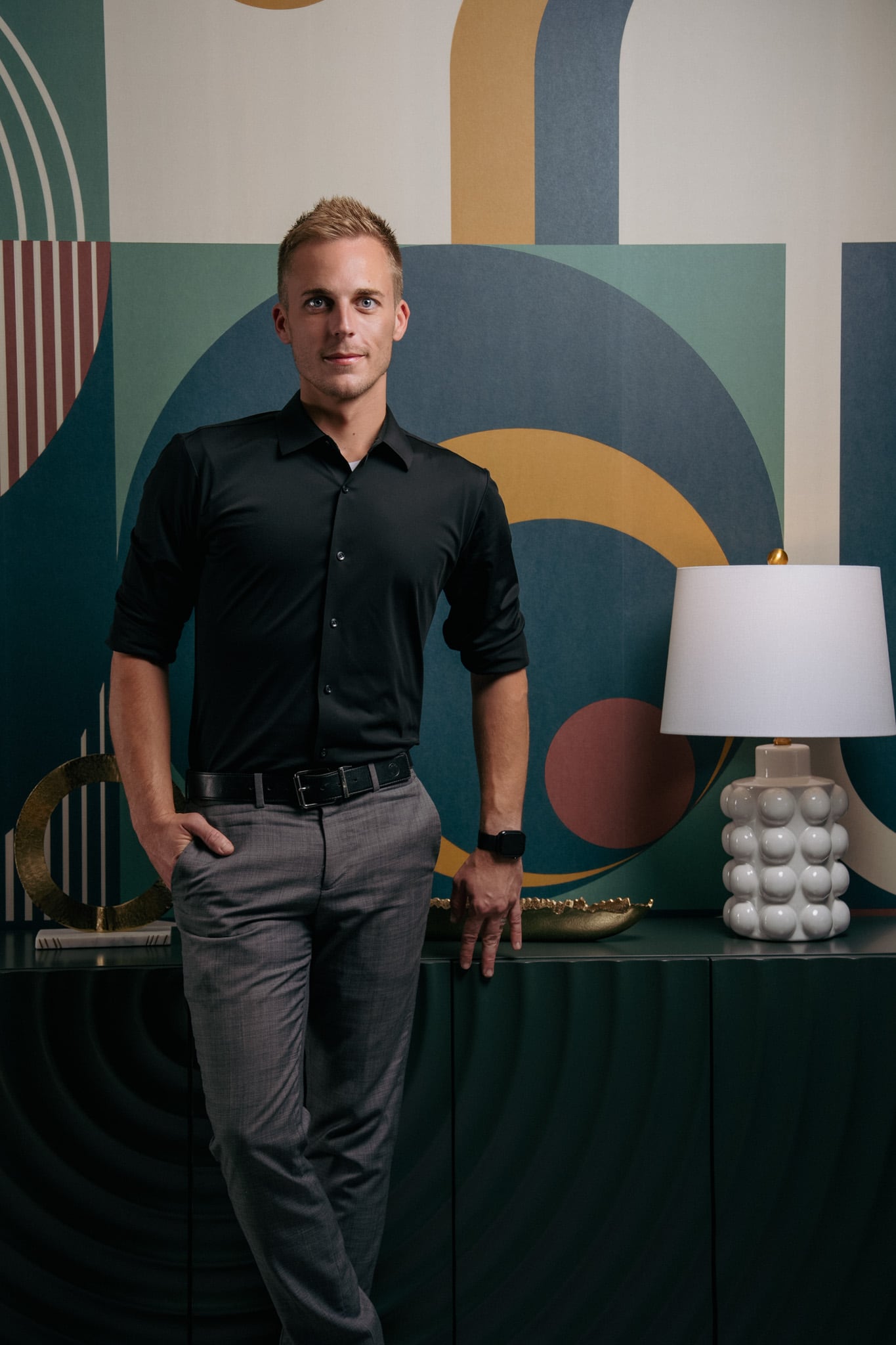 David Weis in a black shirt beside a table lamp, in front of a painted mural in blue, green and orange