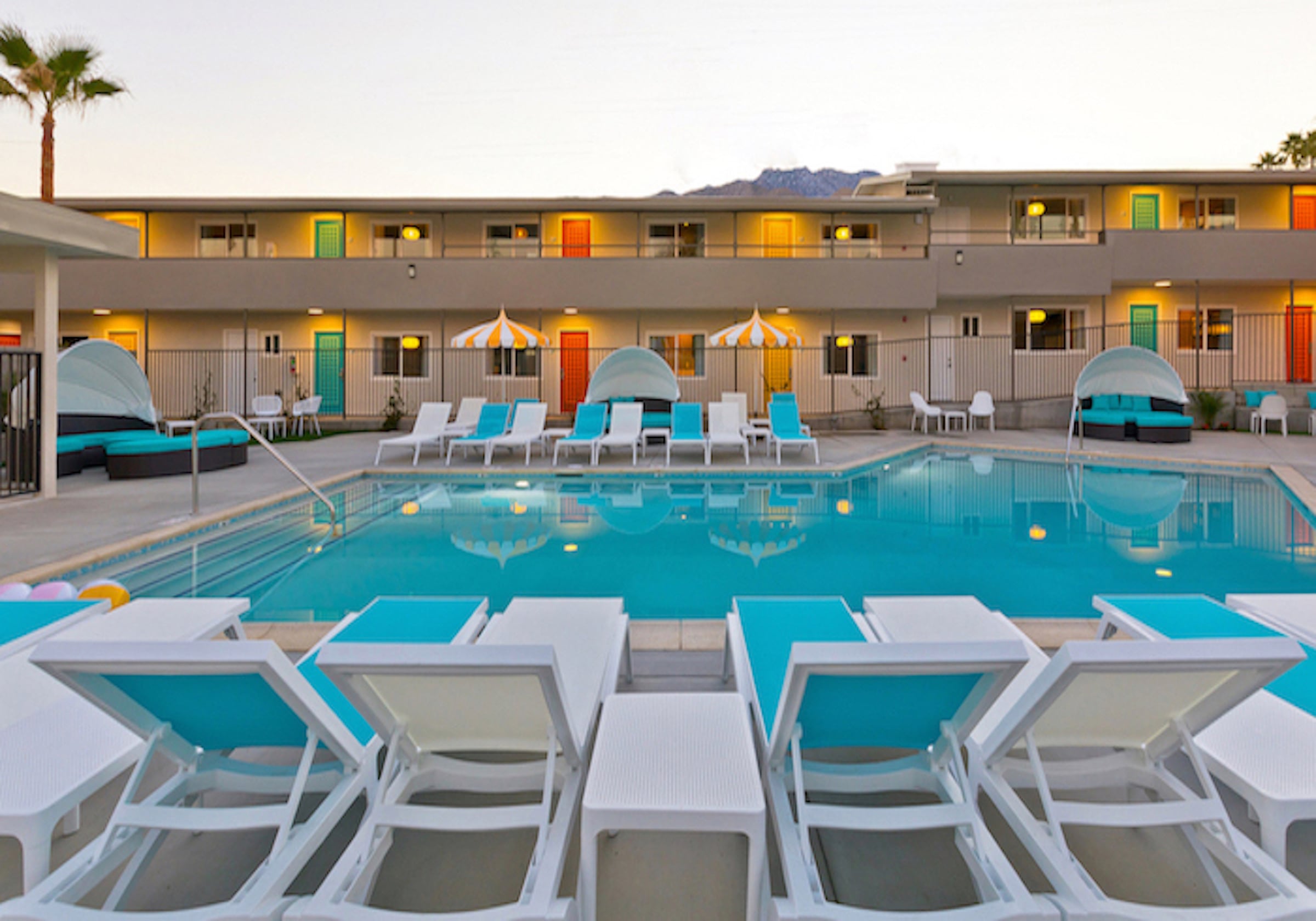 The Cole Hotel pool at dusk: white loungers with turquoise cushions, striped umbrellas and the two-story motor-lodge building with its orange, yellow and green doors lit behind