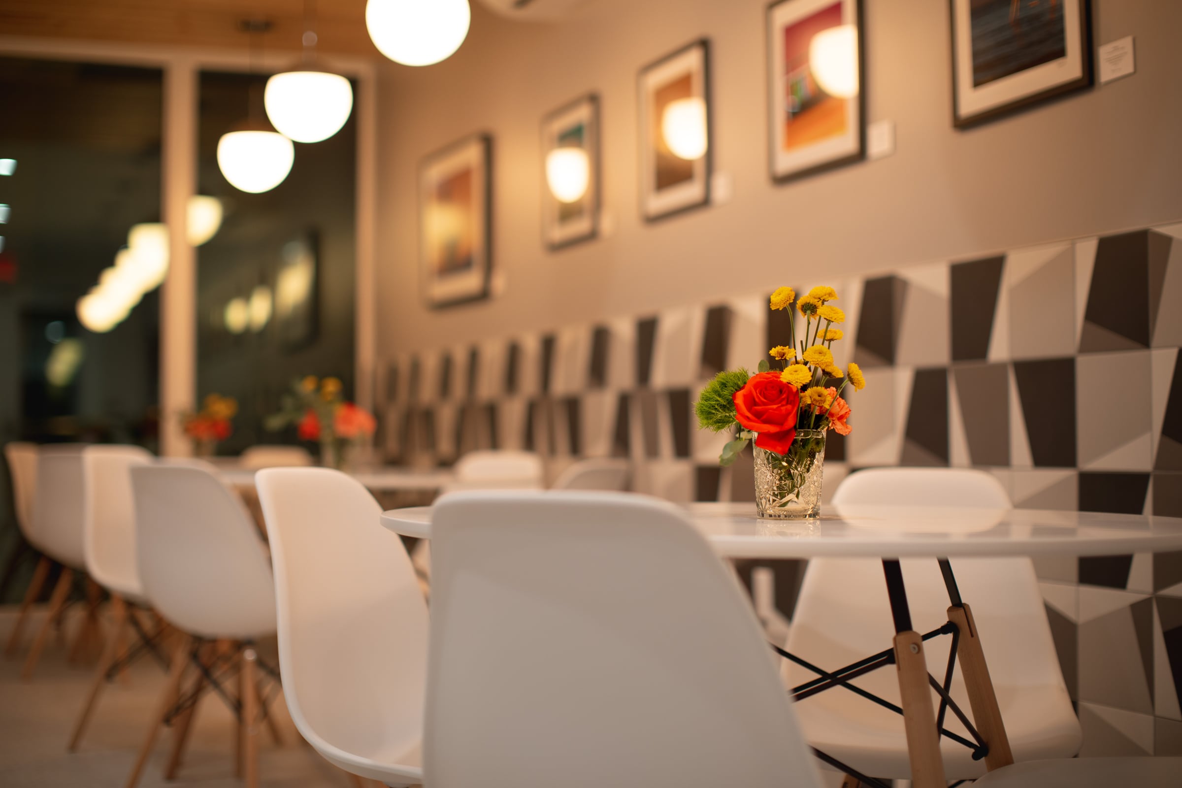 The restaurant at The Cole: white molded chairs at long tables, a grey geometric tiled wall hung with photographs, and a vase of red and yellow flowers