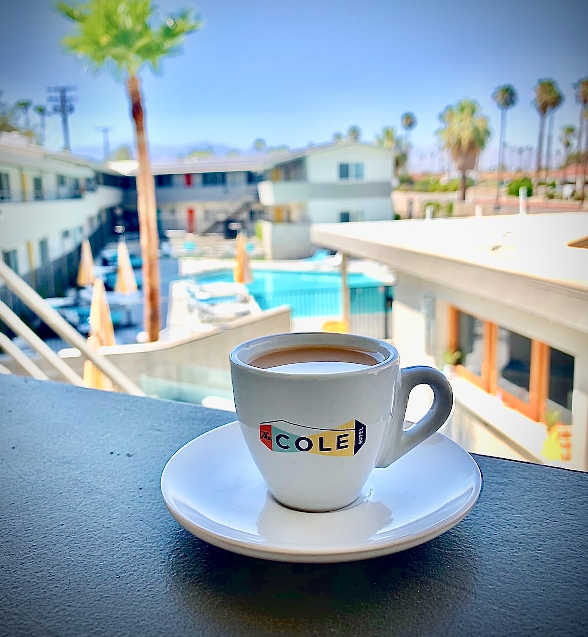 A Cole Hotel branded espresso cup and saucer on a balcony table, with the pool, palms and mountains beyond