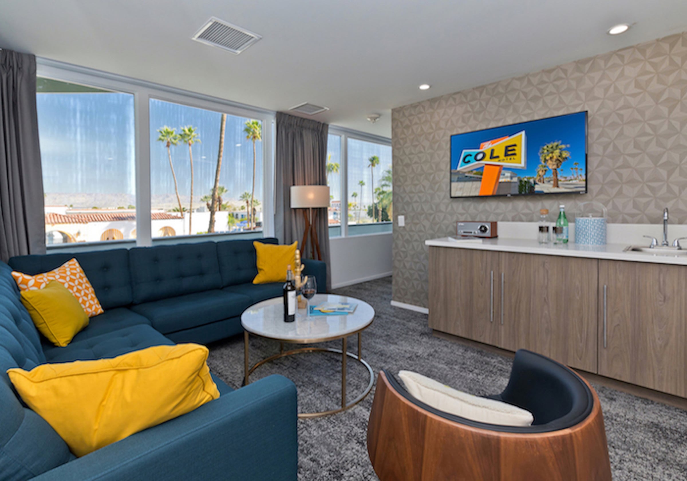 A guest suite at The Cole with a teal sectional and yellow pillows, a walnut barrel chair, a kitchenette and palms and mountains through the corner windows