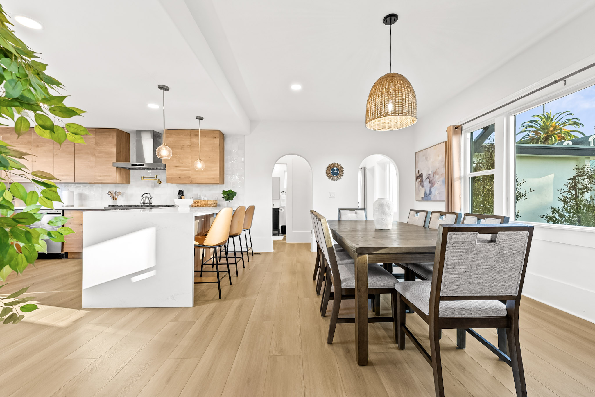 The open kitchen and dining room at Vista Voltaire: oak cabinets, a white waterfall island with tan leather stools, a long wood dining table under a rattan pendant and two arched doorways