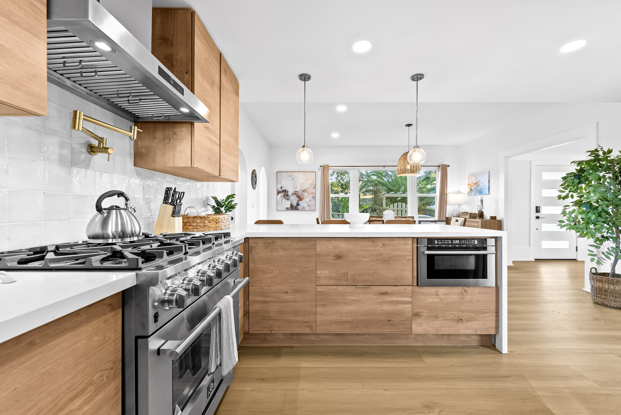 Kitchen detail at Vista Voltaire: a stainless range and hood, a brass pot filler on glossy white tile and oak cabinets, with the front door visible beyond
