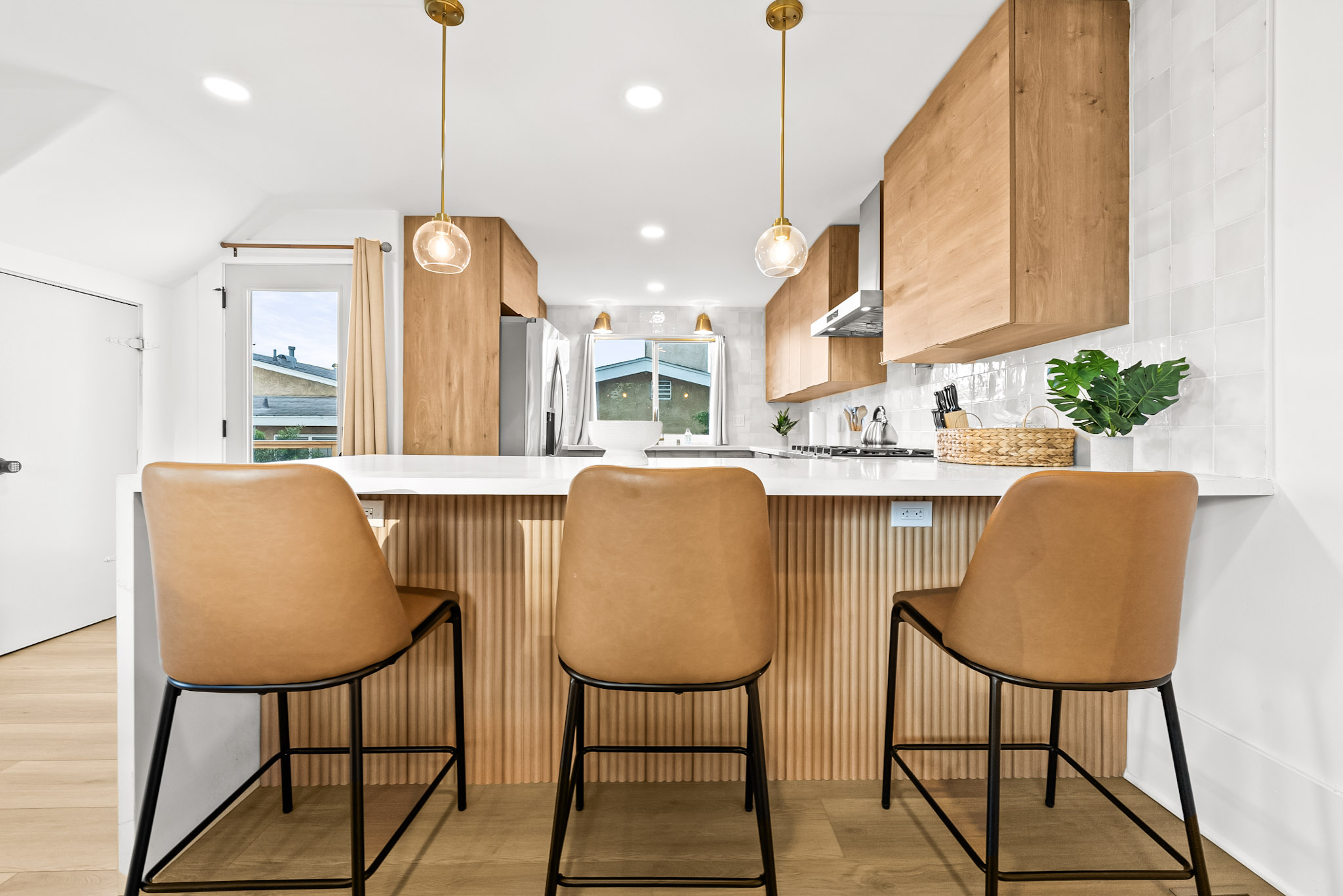Three tan leather stools at the fluted oak breakfast bar in the Vista Voltaire kitchen, under two brass and glass pendants