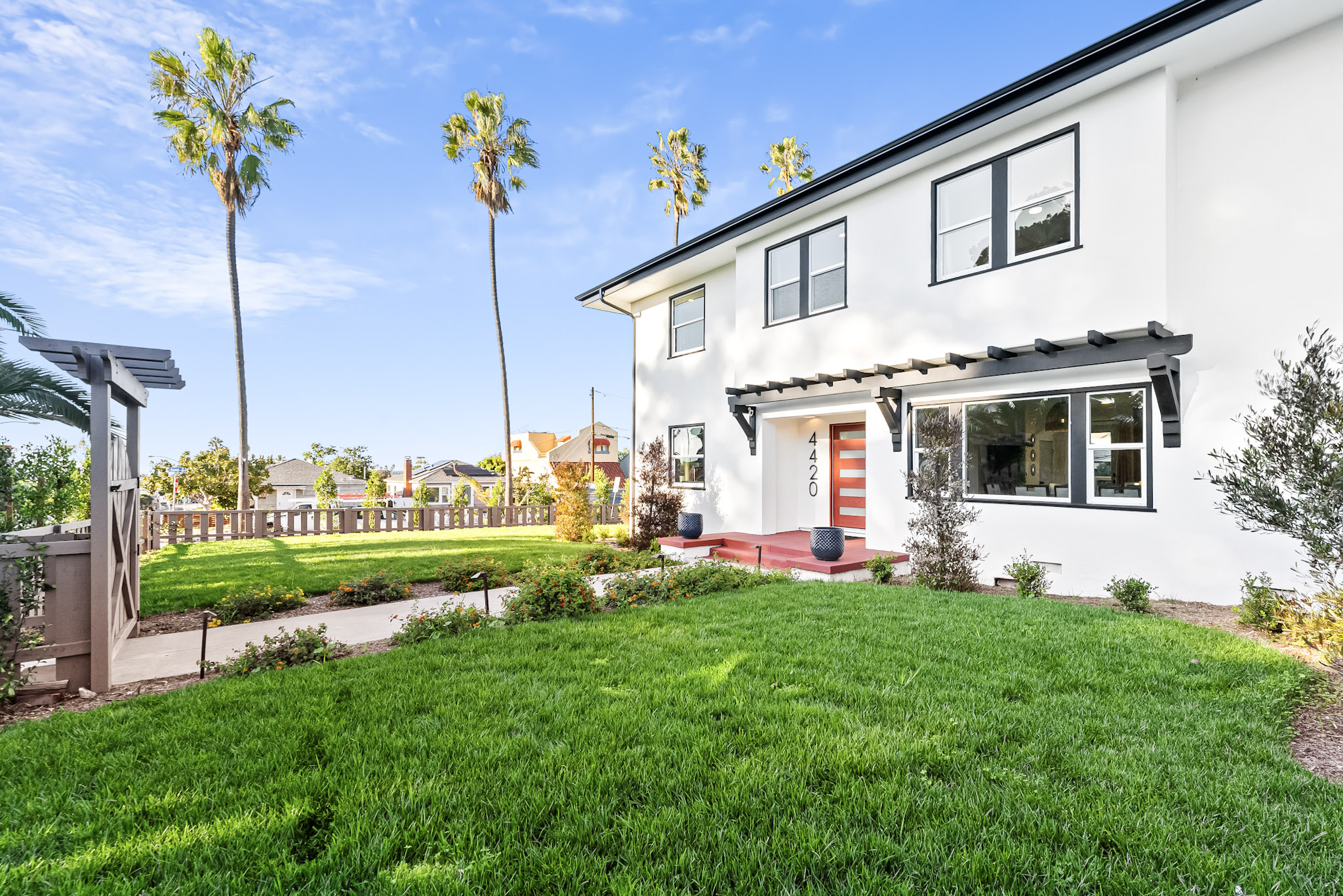 Vista Voltaire from the front corner of the lot: fresh lawn, a gate and picket fence, tall palms and the black pergola over the entry