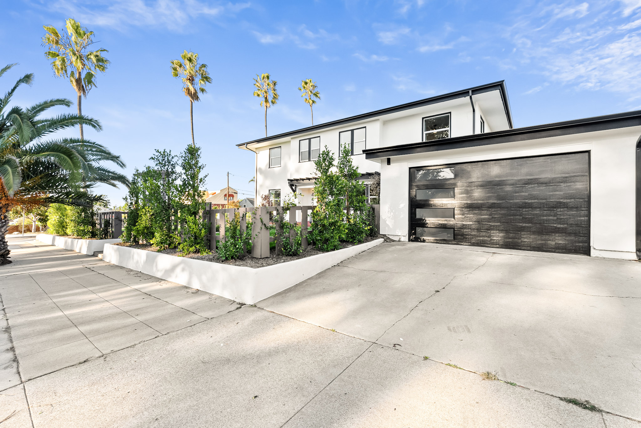 Vista Voltaire from the street, with the driveway, a black garage door, a new hedge inside the fence and palms overhead