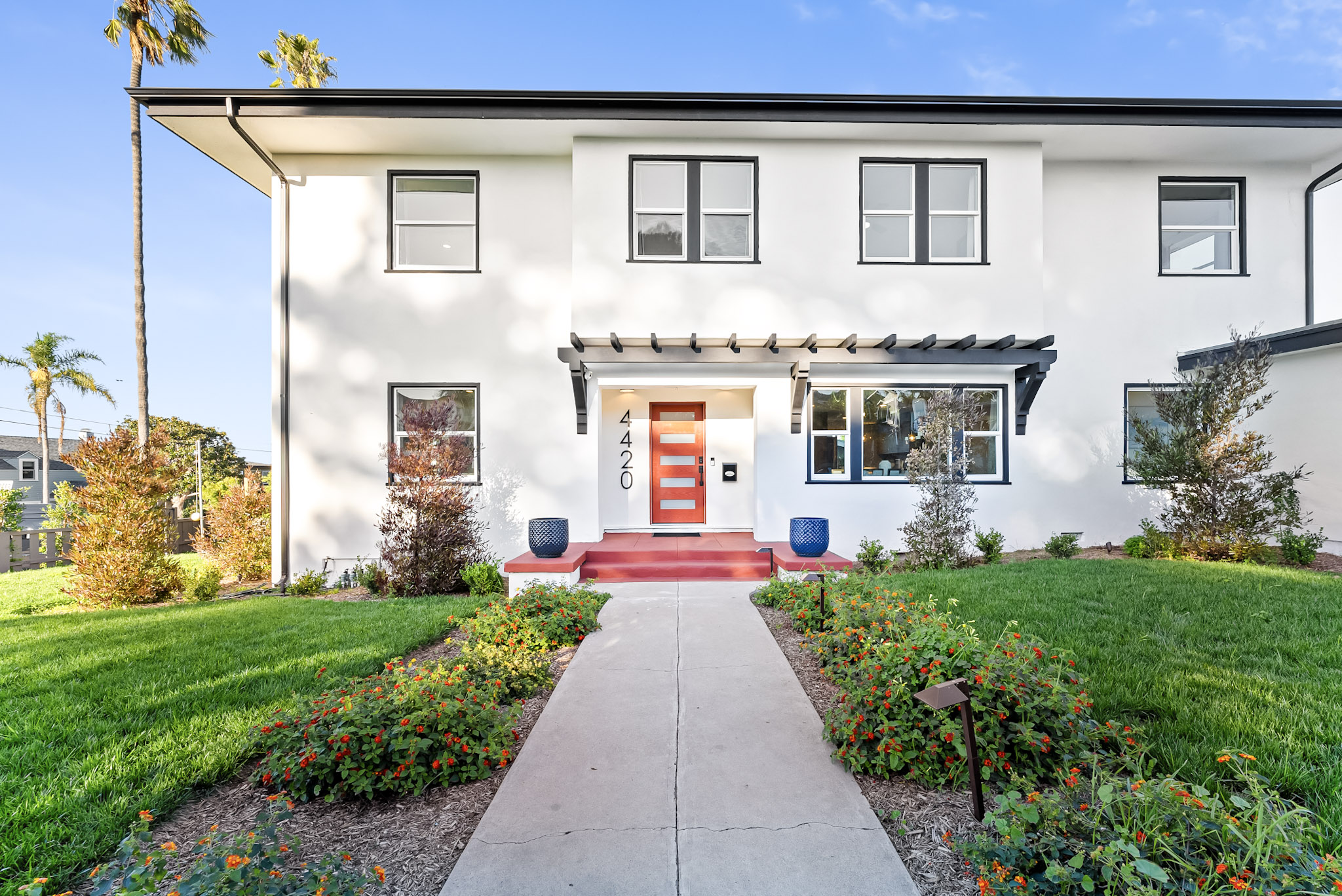 Vista Voltaire after the restoration: a white two-story home in Ocean Beach with black-trimmed windows, a wood front door under a pergola, red steps and a walkway lined with flowering lantana